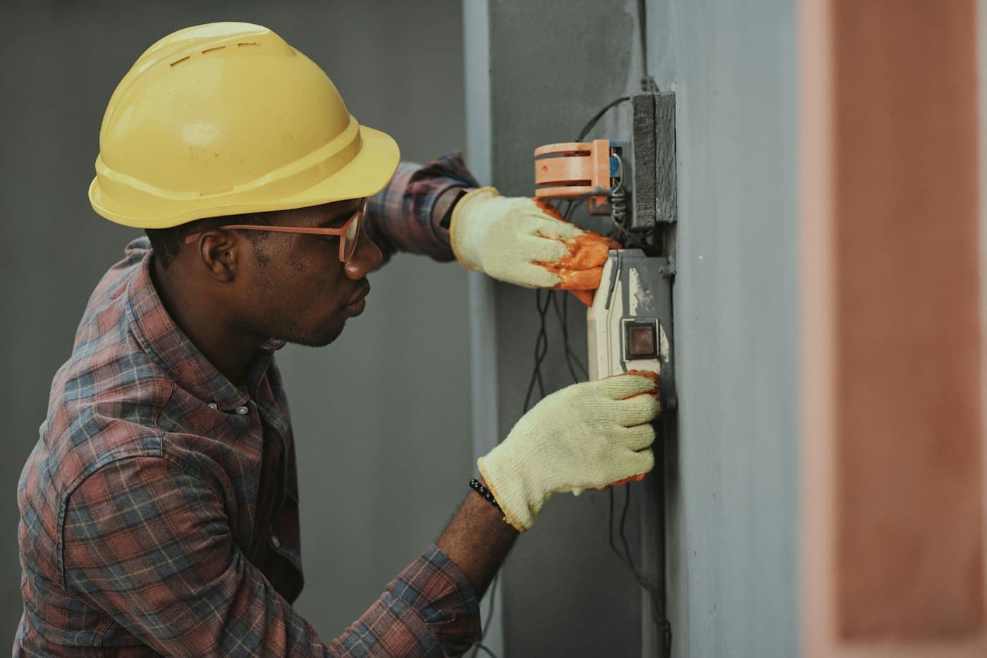 An electrician in a hard hat and gloves works on a meter mounted on a wall