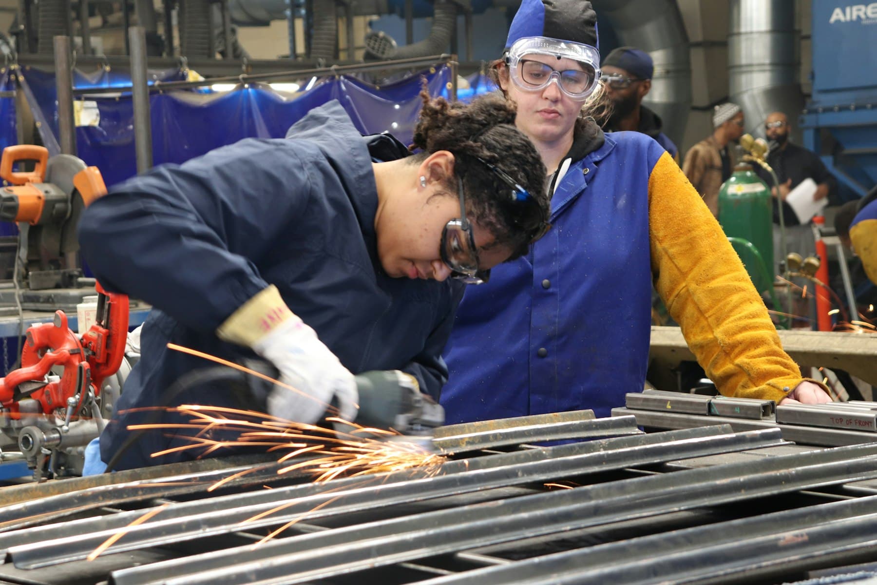 A trainee in safety glasses grinds steel in a training shop while an instructor watches